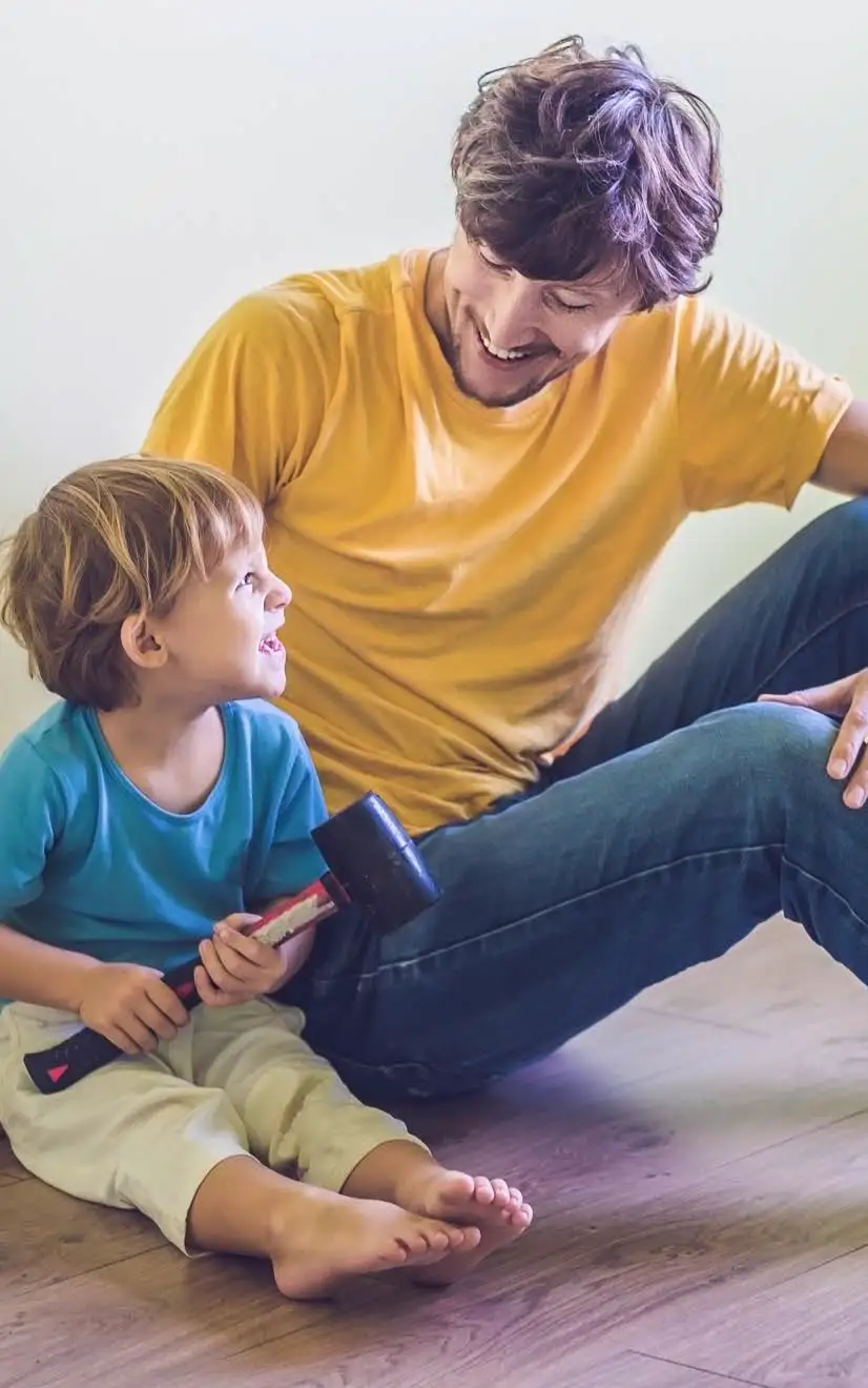 A father and young son. The son holds one of the father's tools, a rubber mallet.